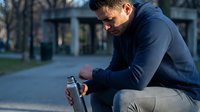 Young athletic Caucasian man in navy hoodie sitting outdoors holding water bottle after workout, urban fitness lifestyle concept for health and wellness.