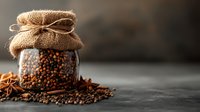 Glass jar filled with aromatic black peppercorns, topped with rustic burlap and twine, scattered spices and star anise on dark surface with moody lighting.