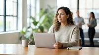 Young woman with curly dark hair and medium skin tone working on laptop at wooden desk in modern office with plants and colleagues in background.