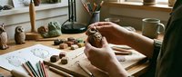 Hands sculpting a clay owl figurine at a wooden craft table with tools, sketches, and ceramic owl statues in a cozy studio.