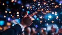 Male hand in business suit pointing upward against blurred festive bokeh lights and confetti background, creating magical celebratory atmosphere at corporate event.