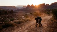 Hiker with backpack trekking through desert landscape at golden hour sunrise, surrounded by red rock formations and sparse vegetation, adventure travel and outdoor exploration.