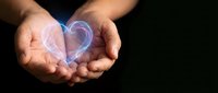 Cupped hands holding glowing translucent heart symbol against dark background representing love, care, protection and emotional support concepts.