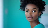 Young African American woman with natural curly hair and confident expression wearing turquoise medical scrubs against blurred hospital background.