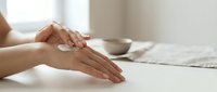 Woman applying moisturizer cream on hands at white table with cosmetic jar in background. Skincare routine and hand care concept for beauty products.