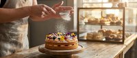 Baker dusting powdered sugar on chocolate cake with edible flowers in warm bakery kitchen with fresh pastries displayed on shelves in background.