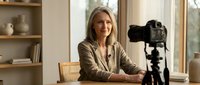Middle-aged woman with blonde hair and fair complexion, wearing a beige cardigan, seated at a desk with a professional camera on tripod, ready for content creation or video recording in a modern home studio setting.