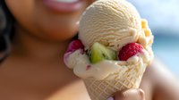 Creamy vanilla ice cream scoop in wafer cone garnished with fresh raspberries and kiwi slices, held against blurred background. Perfect for food advertising.
