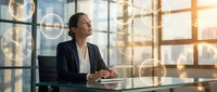 Professional businesswoman in dark suit working at modern office desk with laptop, looking thoughtfully toward bright windows with warm sunlight streaming in.