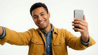 Young man with dark hair and medium brown skin tone wearing mustard yellow jacket over denim shirt taking selfie with smartphone, arms outstretched, smiling at camera against white background for social media and technology concepts.