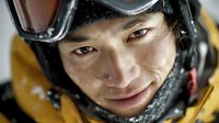Young Asian male mountaineer with protective goggles and winter gear smiling after extreme cold weather expedition in snowy mountain conditions.