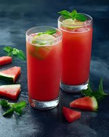 Fresh watermelon juice cocktails garnished with mint leaves and lime slices in tall glasses, served with watermelon pieces on dark background. Refreshing summer drink.