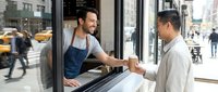 Male barista with dark hair and beard, South Asian ethnicity, wearing denim apron, serving coffee to male customer with light skin tone at urban cafe window counter during daytime.