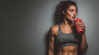 Athletic young woman drinking protein shake after workout in gym setting with dramatic lighting for fitness and nutrition marketing.