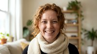 Happy middle-aged Caucasian woman with curly red hair wearing cozy cream turtleneck sweater smiling warmly in bright modern living room with plants.