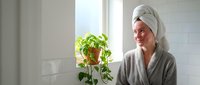 Young Caucasian woman in bathrobe with towel on head holding pothos plant in modern bathroom, morning skincare routine and self-care wellness lifestyle.