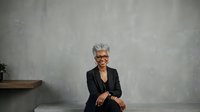 Mature woman with gray hair and glasses wearing black blazer sitting against textured gray wall background for corporate portraits and professional headshots.