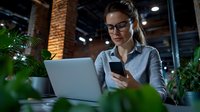 Professional businesswoman with glasses using smartphone and laptop in modern office workspace with brick walls and plants for remote work productivity.