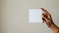 Woman with henna-decorated hand holding blank white card against neutral beige wall. Suitable for greeting cards, invitations, announcements.
