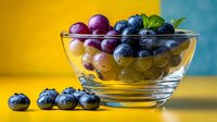Fresh assortment of blueberries, red and green grapes in glass bowl against vibrant yellow and blue background, perfect for food blogs and healthy eating content.