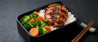 Teriyaki glazed chicken with sesame seeds served over steamed rice with fresh broccoli and carrots in black bento box, ready for lunch or dinner meal.