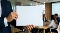 Male professional in dark business suit holding blank white clipboard in modern office meeting room with colleagues in background, suitable for corporate presentations and business communications.