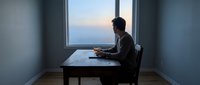 Silhouette of businessman working on laptop at desk by window during sunset, professional remote work setup in modern office space.