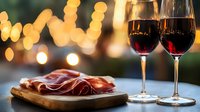 Elegant wine glasses filled with red wine paired with sliced cured meat on wooden board, warm bokeh lights creating romantic dining atmosphere.