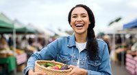 Young woman with long dark hair and medium complexion wearing denim shirt holding fresh sandwich at outdoor farmers market with vendor tents in background.