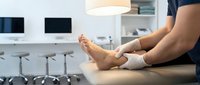 Woman patient sitting on examination table in modern medical office receiving foot ankle treatment from healthcare professional.