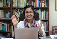 Young woman with headphones waving during video call, sitting at desk with laptop in home office with bookshelf background, wearing white shirt and patterned scarf.