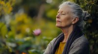 Senior woman with gray hair in cardigan and yellow top meditating peacefully outdoors in garden setting with soft natural lighting and bokeh background.