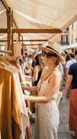 Woman in white linen dress and straw hat browsing clothing at outdoor market stall with fabric canopy, summer shopping scene.