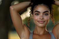 Young mixed-race woman with curly hair in sports bra posing with arms raised outdoors in natural lighting for fitness and wellness lifestyle content.