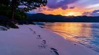 Tranquil beach scene with footprints in white sand along shoreline at sunset, dramatic purple and orange sky reflects in calm water, silhouetted mountains frame horizon.