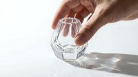 Male hand holding a faceted crystal clear glass on white background, casting rainbow light reflections on surface.