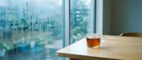 Cozy rainy day scene with a glass cup of hot tea on a wooden table, foggy window with welcome written in condensation, soft natural light.