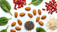 Healthy superfood ingredients on white background including almonds, goji berries, chia seeds, spinach leaves and oats for nutritious meal preparation.