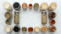Assorted grains, seeds, and legumes in glass containers arranged on marble surface, overhead view for cooking, baking, and healthy eating content.