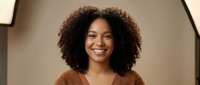 Young African American woman with curly hair smiling warmly in brown sweater against neutral background for professional headshot and portrait use.