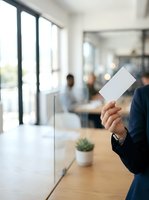 Person in dark business attire holding blank white card in modern office with colleagues and natural light from windows, suitable for corporate branding and business communications.
