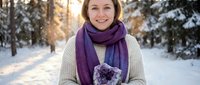 Smiling young Caucasian woman holding amethyst crystal geode in snowy winter forest, wearing cozy sweater and purple scarf, soft sunlight background.