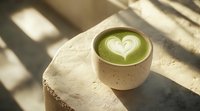 Green matcha latte with heart-shaped foam art in ceramic cup on concrete surface, natural window light casting geometric shadows creates cozy atmosphere.
