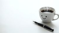 Steaming coffee cup with smiley face and black pen on white surface for morning productivity and positive workspace motivation.