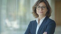 Professional middle-aged Caucasian businesswoman wearing glasses and navy suit, standing with arms crossed in modern office setting with soft natural lighting.