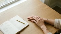 Woman's hands resting on wooden desk next to open notebook with pencil, peaceful workspace moment for planning and reflection in natural light.