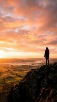 Silhouetted figure standing on rocky mountain peak overlooking vast valley landscape during golden sunset with dramatic orange and yellow clouds, conveying adventure and contemplation.