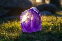 Raw amethyst crystal point on green grass illuminated by golden sunlight, showcasing natural purple hues and crystalline structure in magical outdoor setting.