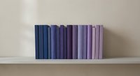 Row of purple and blue books arranged by color gradient on white shelf against neutral wall for home decor and organization concepts.