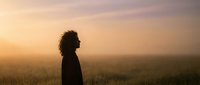 Young woman with curly hair silhouette standing in golden wheat field during sunset, peaceful countryside landscape for wellness and mindfulness concepts.